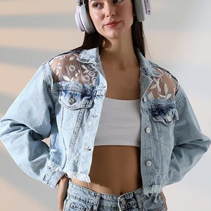 Peut inclure: Une veste en jean bleu clair avec des broderies florales blanches sur des empiècements en maille transparente aux épaules. La veste présente une fermeture boutonnée, deux poches poitrine à rabat et un ourlet effiloché. Portée sur un haut court blanc.