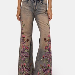 Op de afbeelding: High-waisted flared jeans in een vervaagde blauwe denim wassing met gedetailleerd bloemenborduursel in roze, paarse en groene tinten langs de zijkanten van de pijpen. De broek heeft een klassiek ontwerp met vijf zakken en een knoopsluiting.