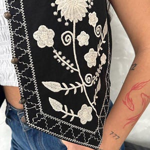 Puede incluir: Un chaleco negro con intrincados bordados florales blancos y un borde decorativo en zigzag a lo largo del frente. La prenda se asegura con una hilera de peque&ntilde;os botones met&aacute;licos oscuros y redondos situados en la tapeta frontal vertical.