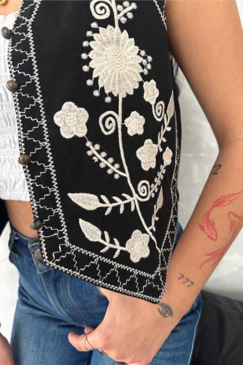 Puede incluir: Un chaleco negro con intrincados bordados florales blancos y un borde decorativo en zigzag a lo largo del frente. La prenda se asegura con una hilera de peque&ntilde;os botones met&aacute;licos oscuros y redondos situados en la tapeta frontal vertical.