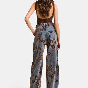 May include: A brown bodysuit paired with wide-leg blue jeans featuring a brown floral pattern. The jeans have a high waist and a relaxed fit. A brown belt is worn at the waist. The model is wearing brown heels.