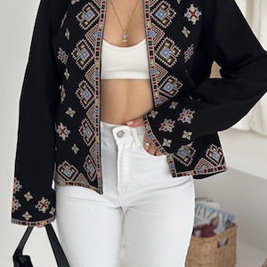 May include: A black long-sleeved jacket featuring intricate multi-colored geometric embroidery along the front opening and cuffs. The jacket is worn over a white cropped top and paired with white denim jeans, accompanied by a black handbag.