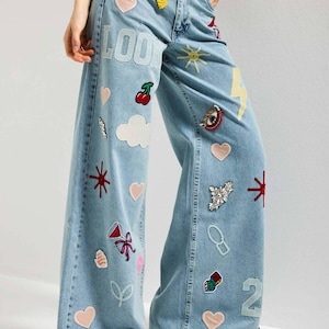 May include: Light blue denim wide-leg jeans featuring various embroidered patches including hearts, a yellow lightning bolt, a sun, cherries, a red eye, dice, and the word LOOK in white letters. The pants have a high-waisted fit and a loose silhouette.