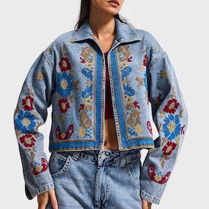 Puede incluir: Chaqueta vaquera azul claro con bordados florales coloridos en rojo, azul y dorado. La chaqueta tiene cuello, ribete azul y corte cropped. Mangas largas, combinada con vaqueros a juego.