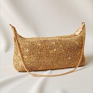 Peut inclure: Un sac à main rectangulaire doré doté d'une texture en maille dense et scintillante. Le sac présente une ouverture supérieure structurée avec une fine bandoulière en chaîne dorée assortie fixée des deux côtés, offrant un aspect métallique uniforme.