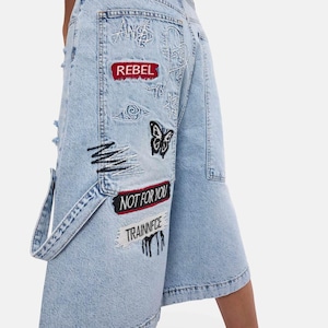Può includere: Culotte in denim azzurro con varie toppe ricamate, tra cui un rettangolo rosso con la scritta REBEL, una farfalla nera, un rettangolo nero con la scritta NOT FOR YOU e una toppa bianca con la scritta TRAINNECE in nero.
