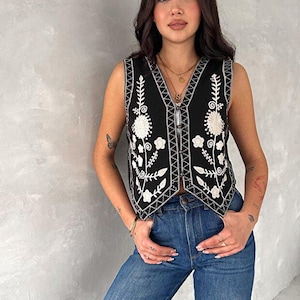 Puede incluir: Un chaleco negro sin mangas con bordados florales blancos en los paneles frontales y escote en V. La prenda tiene un cierre frontal de botones y un dobladillo en punta, combinado con unos vaqueros azules de dise&ntilde;o cl&aacute;sico de cinco bolsillos.