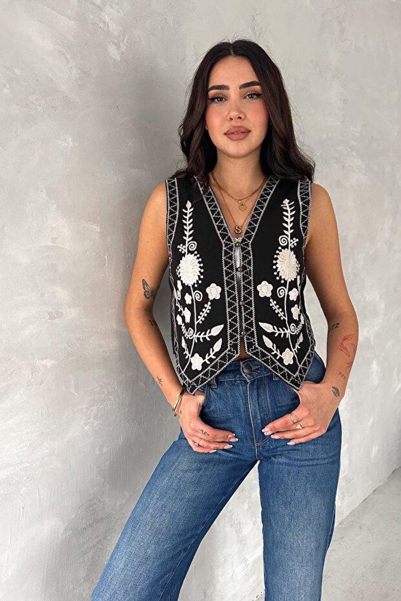 Puede incluir: Un chaleco negro sin mangas con bordados florales blancos en los paneles frontales y escote en V. La prenda tiene un cierre frontal de botones y un dobladillo en punta, combinado con unos vaqueros azules de dise&ntilde;o cl&aacute;sico de cinco bolsillos.