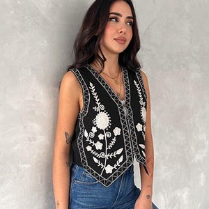 Puede incluir: Un chaleco negro sin mangas con bordados florales blancos y un patr&oacute;n geom&eacute;trico en los bordes del cuello en V y el dobladillo. La prenda cuenta con un cierre frontal de botones y se combina con pantalones vaqueros azules para un conjunto.