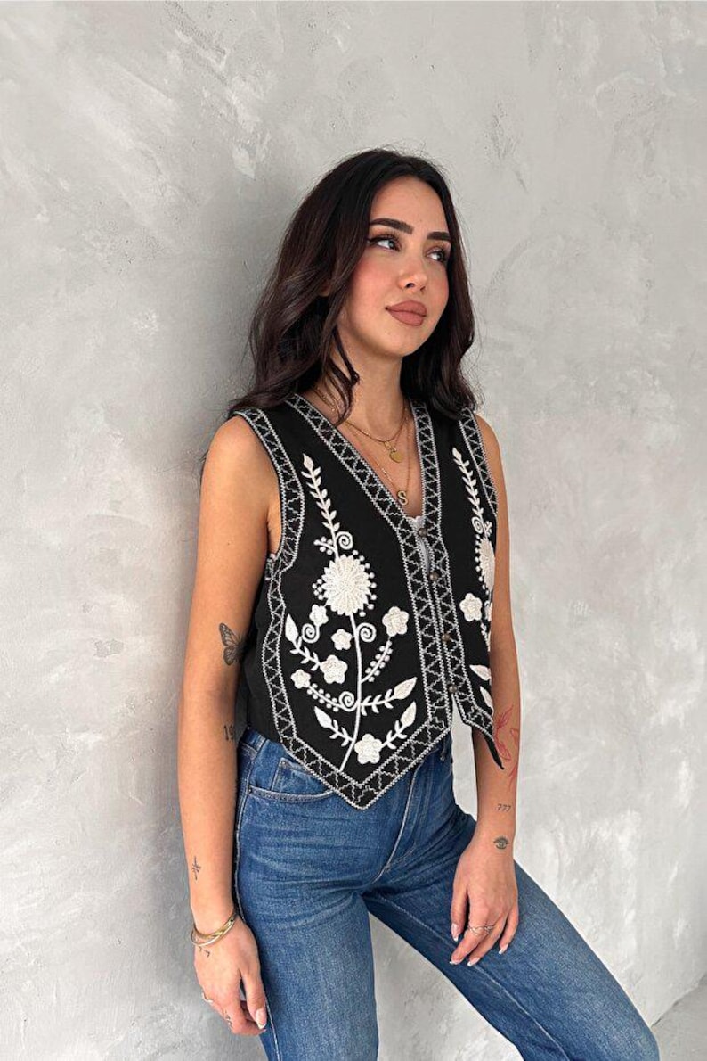 Puede incluir: Un chaleco negro sin mangas con bordados florales blancos y un patr&oacute;n geom&eacute;trico en los bordes del cuello en V y el dobladillo. La prenda cuenta con un cierre frontal de botones y se combina con pantalones vaqueros azules para un conjunto.
