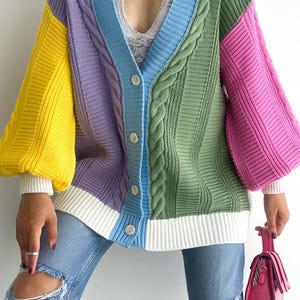 Pode incluir: Um cardigã de malha trançada com blocos de cores, carcela em V azul-claro com botões brancos, uma manga amarela, um painel roxo, um painel verde e uma manga rosa. A peça possui punhos e bainha canelados na cor branca.