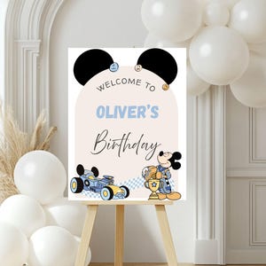 Op de afbeelding: Een welkomstbord voor een verjaardagsfeestje met Mickey Mouse. Op het bord staat "Welkom op Oliver's Verjaardag" in blauwe en zwarte letters. Mickey Mouse wordt afgebeeld met een trofee en een raceauto. Het bord wordt weergegeven op een houten ezel, met witte ballonnen op de achtergrond.