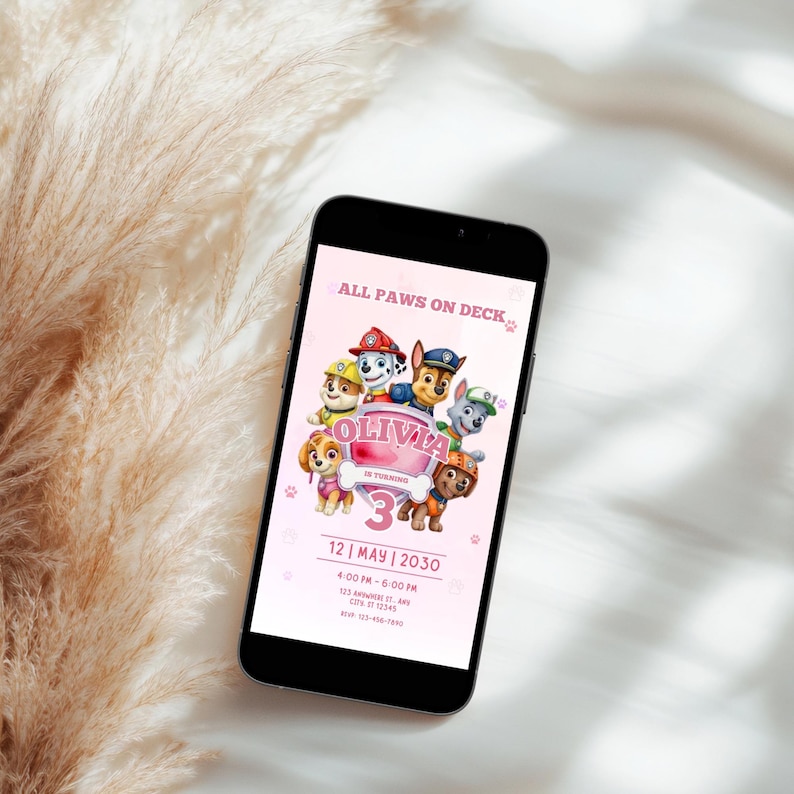 May include: A smartphone displays a pink birthday invitation with the text "ALL PAWS ON DECK" and "OLIVIA 3". The invitation features cartoon characters and event details: date, time, and location. The phone is set against a soft, neutral background.