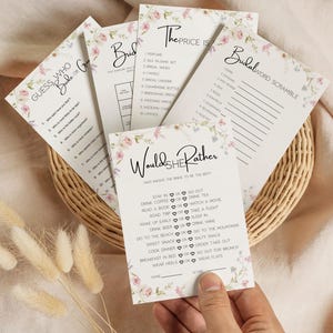Puede incluir: Un conjunto de tarjetas de juegos para una despedida de soltera con detalles florales, incluyendo "Adivina Quién es la Novia o el Novio", "El Precio Justo" y "Sopa de Letras Nupcial". Las tarjetas están dispuestas en una cesta de mimbre, con una mano sosteniendo una tarjeta.