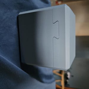May include: A light gray, box-shaped object with a unique interlocking design on one side. The object has a matte finish and appears to be made of a solid material. The box is held against a dark blue fabric.