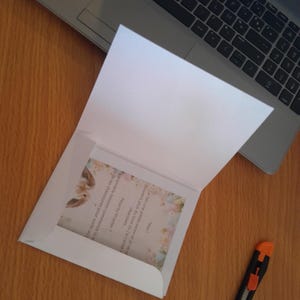 Puede incluir: Un sobre blanco abierto que revela una tarjeta con un diseño floral y texto en francés. El conjunto está sobre una superficie de madera, junto a un portátil y un cúter.