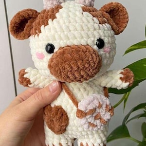 Puede incluir: Un peluche de vaca hecho a mano con parches marrones y blancos, ojos negros y mejillas rosadas. La vaca tiene una pequeña bolsa rosa y blanca y está hecha de hilo suave y texturizado. Un animal de peluche lindo y tierno.