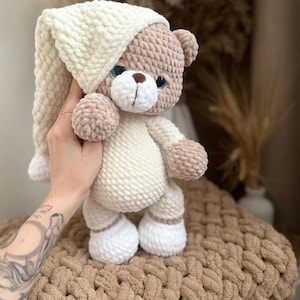 Op de afbeelding: Een handgemaakte gehaakte teddybeer met een crèmekleurige nachtmuts en pyjama. De beer heeft een lichtbruin gezicht, armen en benen, met blauwe ogen en een bruine neus. De beer staat op een bruin, getextureerd oppervlak.