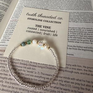 May include: A beaded bracelet with clear beads and gold accents, featuring green, white, and pearl-like beads. The bracelet rests on an open book with the text "truth threaded co." and "THE VINE".