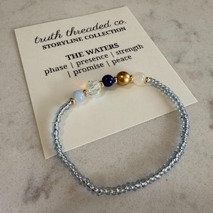 May include: A beaded bracelet with light blue beads and gold accents. The bracelet features a cluster of beads in blue, clear, gold, and white. A card behind the bracelet reads "truth threaded co." and "THE WATERS".