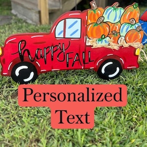 May include: A red wooden truck cutout featuring black wheels with white accents and a truck bed filled with orange, green, and blue pumpkins. The side of the truck is painted with the black text "happy FALL" in a mix of script and block lettering.