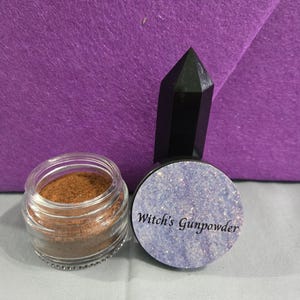 May include: A small glass jar filled with brown powder, a black crystal, and a container with the text "Witch's Gunpowder" on a blue background. The items are set against a purple backdrop.