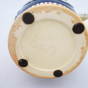 May include: The base of a ceramic beer stein with a white glaze and four black rubber feet. The stein features a blue and white painted design.