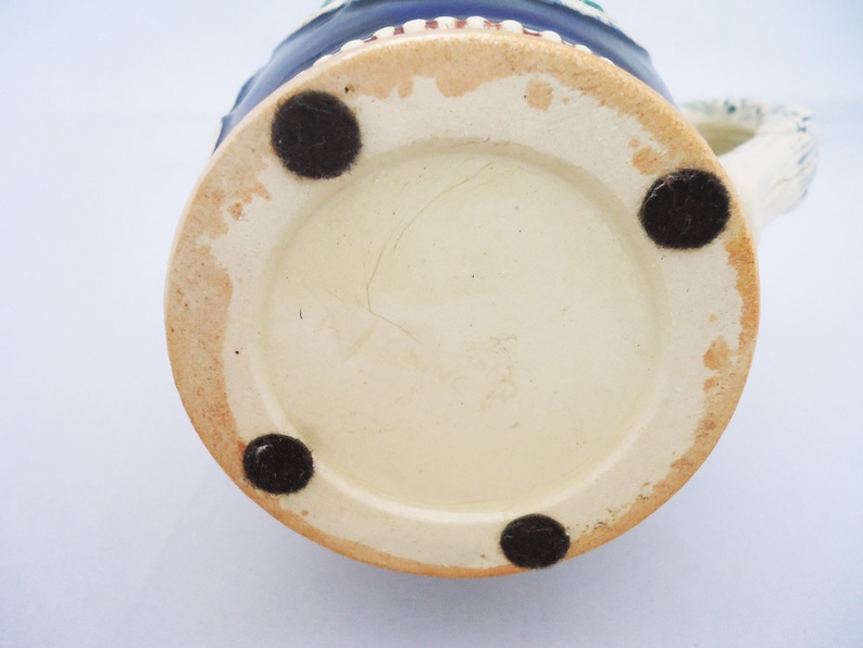 May include: The base of a ceramic beer stein with a white glaze and four black rubber feet. The stein features a blue and white painted design.