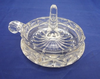 Op de afbeelding: Een glazen ringhouder van helder glas met een decoratief ontwerp. De houder heeft een verhoogde middelste paal om een ring vast te houden en een klein handvat aan de zijkant.