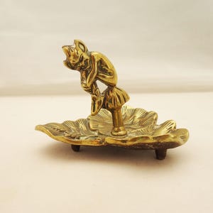 May include: A small, brass figurine of a seated, elf-like creature atop a leaf-shaped dish. The figurine is detailed with a textured surface and rests on three small feet. The overall color is a warm, antique gold.