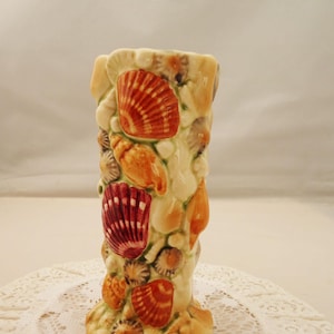 May include: A ceramic vase adorned with assorted seashells in orange, red, and cream hues. The vase is cylindrical with a textured surface, evoking a coastal or nautical aesthetic.
