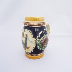 May include: A blue and white ceramic beer stein with a handle. The stein features a scene of a building with a spire and a mountain range. The text "WIEN" is visible on the stein.