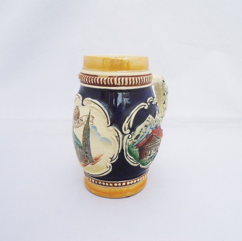May include: A blue and white ceramic beer stein with a handle. The stein features a scene of a building with a spire and a mountain range. The text "WIEN" is visible on the stein.
