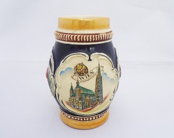 Op de afbeelding: Een blauwe en witte keramische bierkan met een afbeelding van de Stephansdom in Wenen, Oostenrijk. De kan heeft een gouden rand en een bruine handgreep.