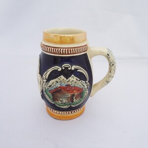 May include: A small blue and white ceramic beer stein featuring a scene of a house in the mountains. The stein has a gold rim and a handle.