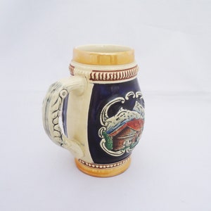 May include: A ceramic beer stein with a blue and white design featuring a mountain scene with a house. The stein has a gold rim and a handle shaped like a twisted rope.