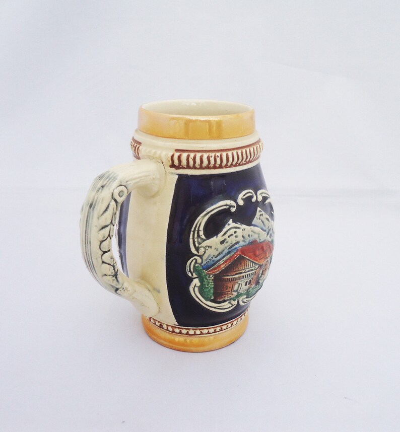 May include: A ceramic beer stein with a blue and white design featuring a mountain scene with a house. The stein has a gold rim and a handle shaped like a twisted rope.