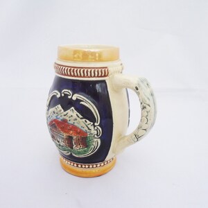 May include: A ceramic beer stein with a blue and white design featuring a mountain scene and a small house. The handle is sculpted with a white design.