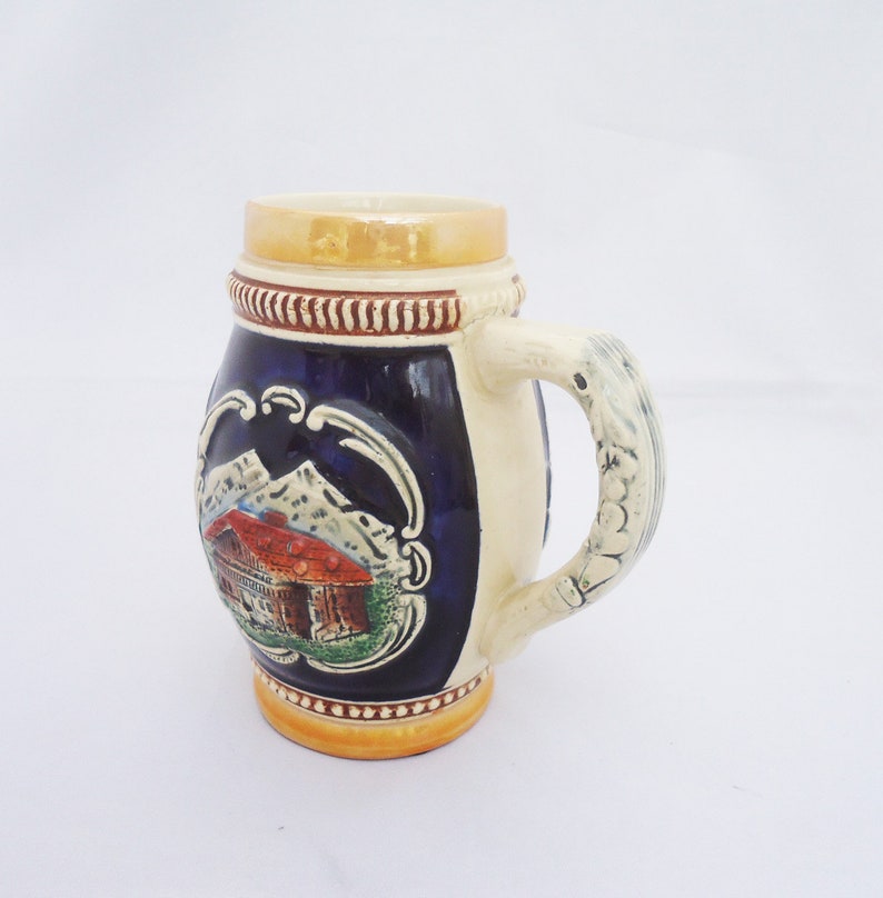 May include: A ceramic beer stein with a blue and white design featuring a mountain scene and a small house. The handle is sculpted with a white design.