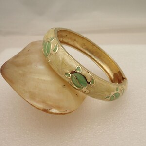 May include: A gold bangle bracelet with a white enamel base and green turtle designs. The bracelet is on a white surface.