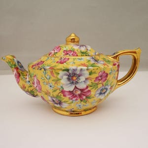 Op de afbeelding: Een gele keramische theepot met een levendig bloemenpatroon in roze, witte en paarse tinten. De theepot heeft een bijpassend deksel met een gouden knop, en is afgewerkt met een goudkleurig handvat, een tuit en een gouden voetrand.