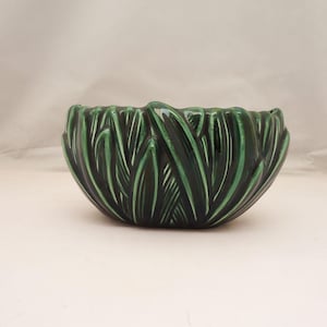 May include: A dark green ceramic bowl with a textured, leaf-like design. The bowl has a glossy finish and a wide, open shape. The exterior features a repeating pattern of vertical, stylized leaves.