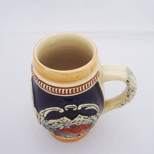 May include: A blue and white ceramic beer stein with a mountain scene and a brown handle.