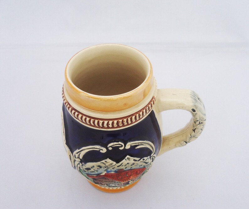 May include: A blue and white ceramic beer stein with a mountain scene and a brown handle.