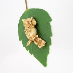 May include: A gold-colored owl brooch with intricate details, perched on a green leaf.
