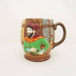 May include: A brown ceramic mug with a 3D design of a man in a green and red costume. The mug has a handle and the words "TOLE WITH WIT OR STEEL" on the bottom.