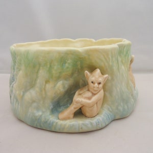 Könnte beinhalten: Ein Keramik-Pflanztopf in Form eines Baumstumpfes mit zwei kleinen, fantasievollen Figuren, die auf der Basis sitzen. Der Topf ist in Grün-, Blau- und Gelbtönen bemalt.