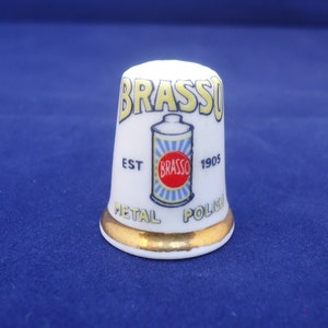 Può includere: Ditale in ceramica bianca con bordo dorato. Il ditale presenta una pubblicità vintage per Brasso Metal Polish. La pubblicità mostra una lattina di Brasso con il testo "Brasso Est 1905 Metal Polish".