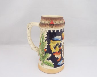 Op de afbeelding: Een keramische bierkan met een blauw en wit bloemenmotief. De kan heeft een handvat en een verhoogde rand. De kan is versierd met een kasteel en andere details.