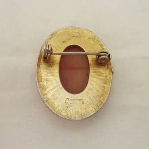 May include: Gold tone oval brooch with a pink cameo set in the center. The back of the brooch has a pin and a clasp. The brooch is marked "MCO."